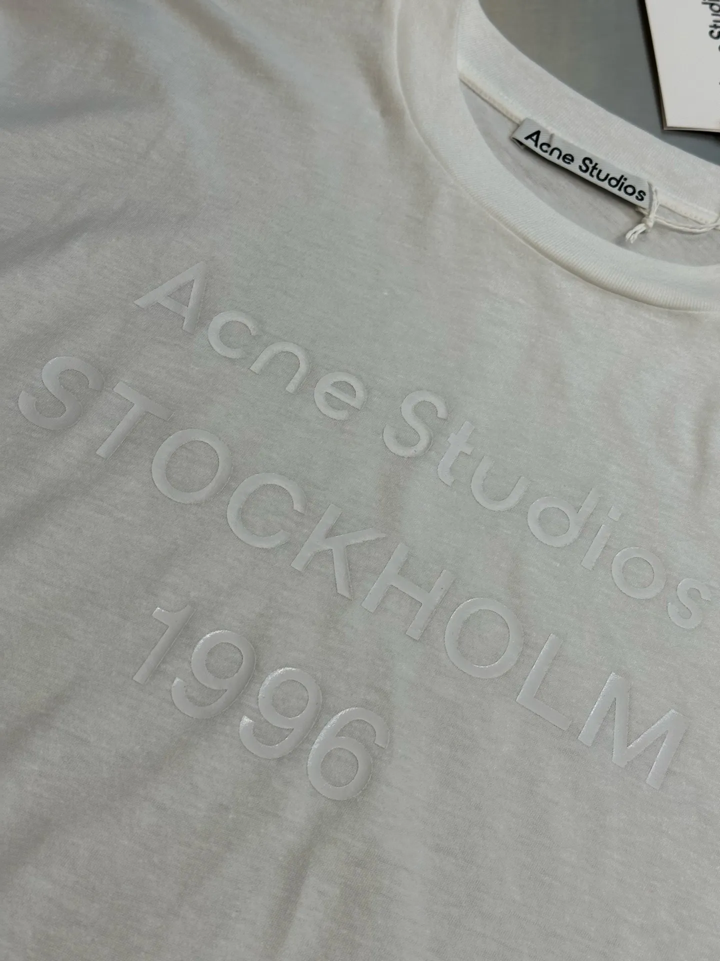 Ac Original White Letter Round Neck Long Sleeve The fabric is made of 32-count cotton and linen blended plain knitting, with a ratio of 85% cotton and 15% linen fiber. The weight is only 145g/m²; the ribbed neckline is made of Same Style cotton and linen blended yarn, 145g/m², to ensure a unified texture of the entire piece. The collision of the natural rough base and the industrial spraying process creates a natural irregular texture and an extremely light and breathable wearing experience. Cotton provides a skin-friendly and soft foundation, while linen introduces natural roughness and knotted particles, giving the fabric surface an irregular texture. The addition of hemp fiber makes the fabric have excellent moisture absorption and perspiration properties, while also bringing a natural micro-wrinkle texture and dry touch. After the fabric is piece-dyed, the dye evenly penetrates into the fiber, showing full color fastness and soft matte color, providing a uniform base for subsequent inkjet printing. The printing uses a double-layer printing process of environmentally friendly elastic glue and varnish, which takes into account the concept of environmental protection, wearing comfort and visual expression. The bottom layer uses environmentally friendly elastic glue, with water-based environmentally friendly resin as the base material. The color paste is scraped onto the surface of the fabric through a high-mesh screen. After drying and solidifying at high temperature, a soft and highly elastic continuous adhesive film is formed. The printed film can stretch and retract with the fabric. Even if it is covered with a large area of ​​pattern, it is still breathable and does not feel tight when wearing it. The surface layer is accurately overprinted with transparent varnish, creating a high-gloss effect in specific areas. This layer of varnish not only significantly improves the color contrast of the pattern, but also creates a local wet and bright visual and tactile contrast, making ordinary printing instantly gain three-dimensional layers and glossy texture. At the same time, the smooth surface layer also enhances Wear-Resistant and wash-resistant properties. This T-shirt is defined as a casual loose cut, unisex style, and hip length. The light fabric slides smoothly along the body contours, forming natural and soft folds at the shoulders and waist. The shoulder line falls naturally, the bust and hem circumferences are similar, and it presents a straight silhouette, without deliberately distinguishing between male and female body shapes. The hip-length design utilizes the elongated body and the extremely light drape of the fabric to create lazy and flowing lines. The ribbed neckline, cuffs and hem are all made of Same Style cotton and linen blended yarn, which maintains a unified overall texture and is loose yet exquisite in details. size：xs s m l